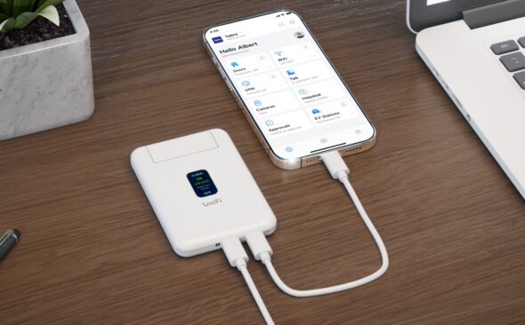 White UniFi Travel Router on a wooden desk plugged into a smartphone by USB-C next to a laptop