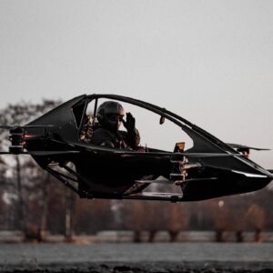 Black YIVTOL S-Zero single-seat eVTOL hovering a few feet off the ground with a helmeted pilot waving