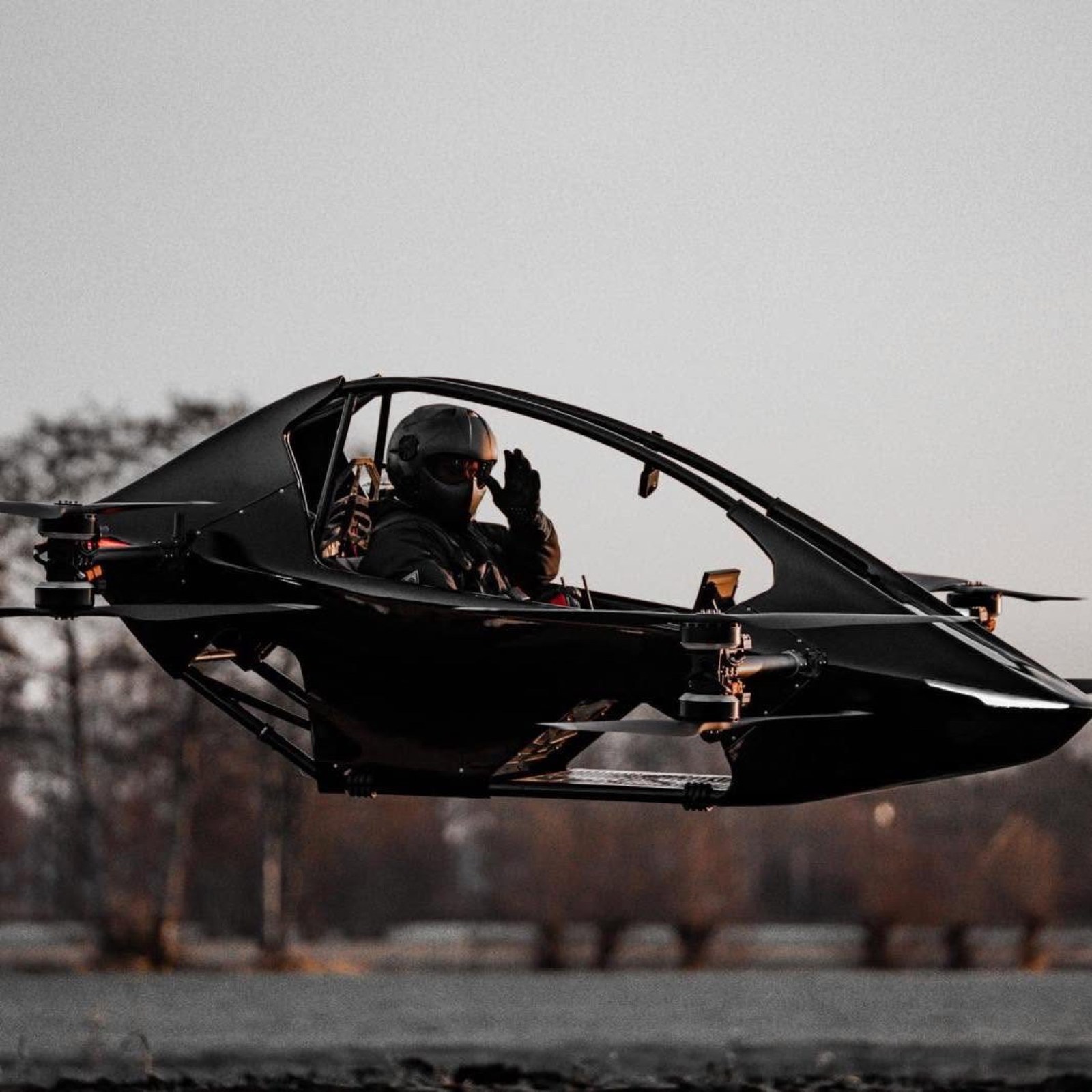 Black YIVTOL S-Zero single-seat eVTOL hovering a few feet off the ground with a helmeted pilot waving