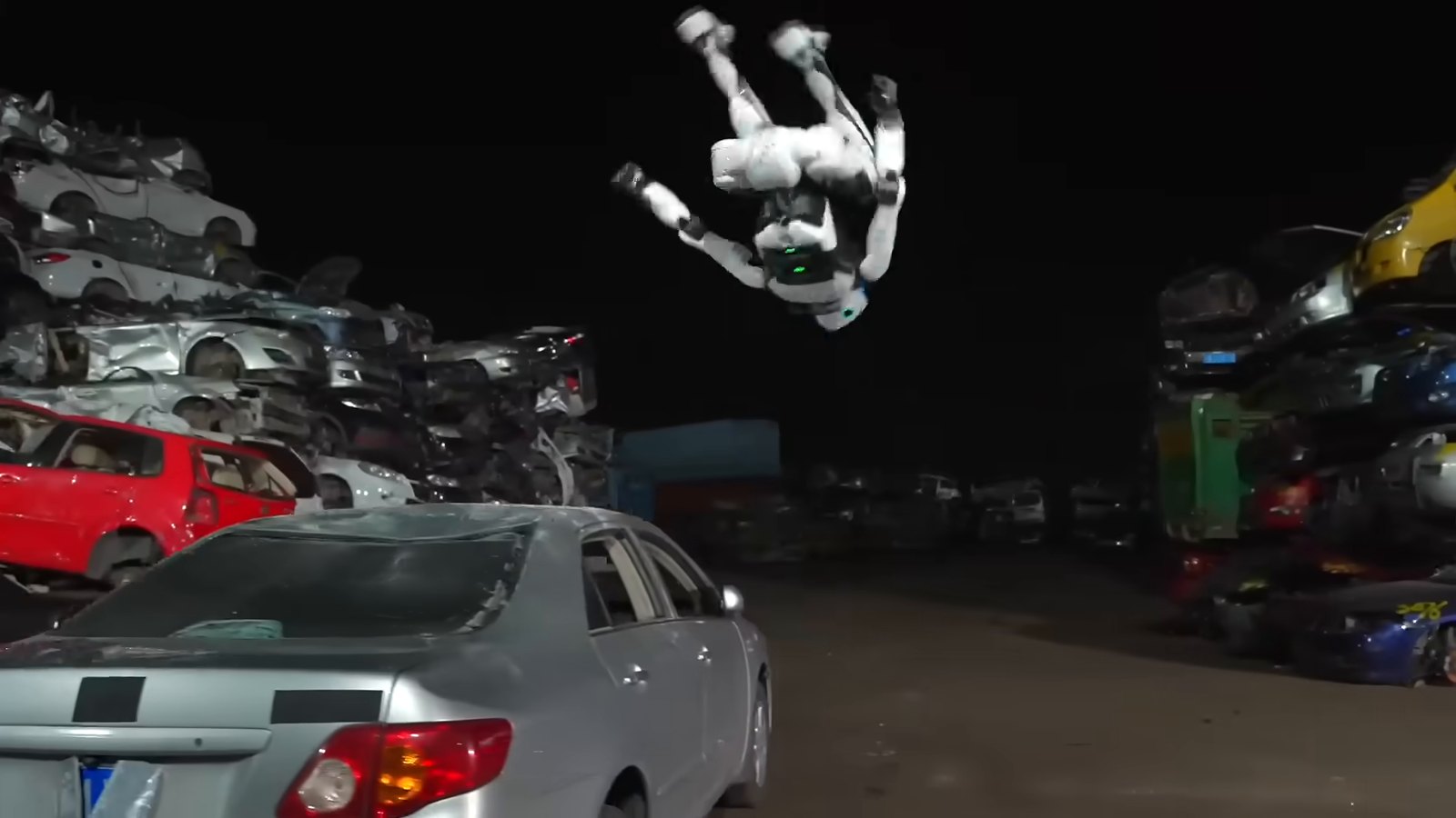 Unitree H2-X robot doing a backflip above a car in a junkyard full of stacked wrecks