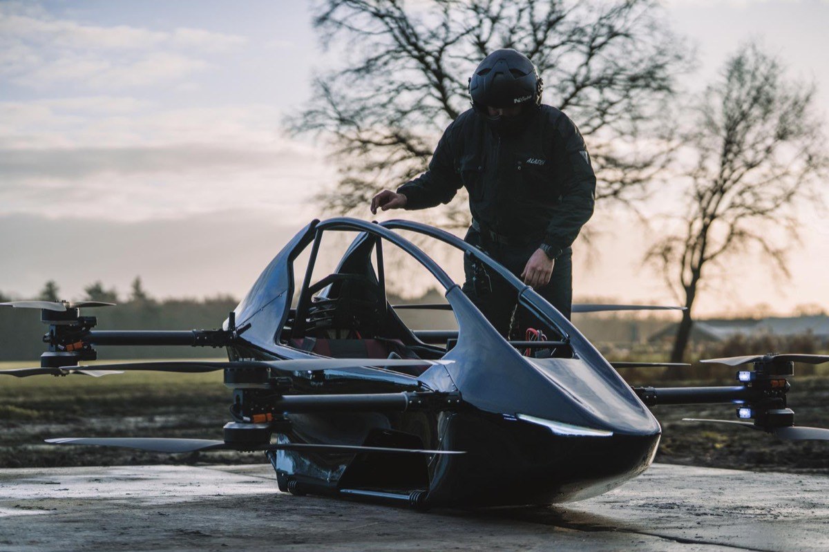Pilot in a black helmet climbing into the open cockpit of a dark blue YIVTOL S-Zero parked on wet pavement