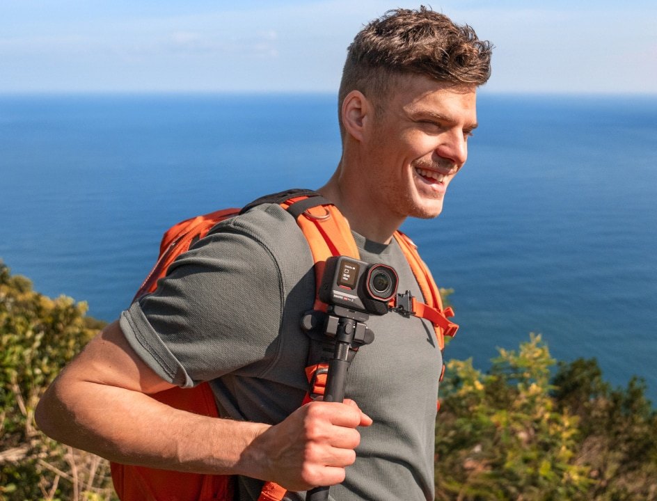 Smiling hiker holding an Insta360 Ace Pro 2 on a handheld grip on a cliff above the ocean