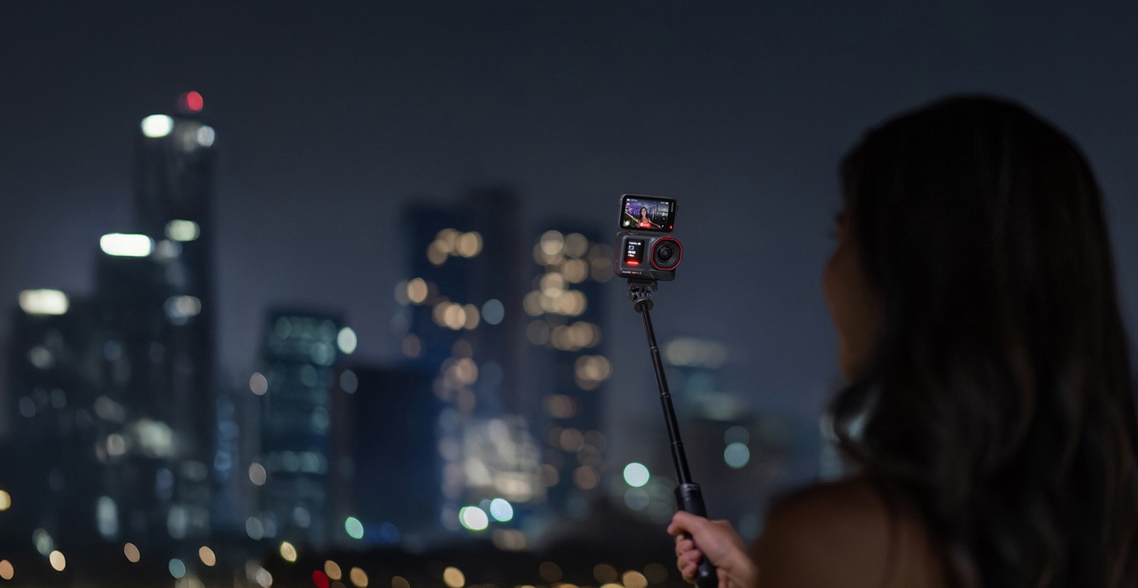 Woman filming herself at night with an Insta360 Ace Pro 2 on a selfie stick in front of a lit city skyline