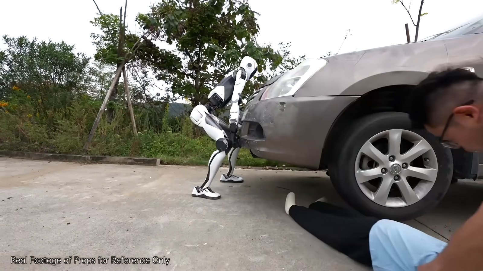 Unitree H2-X lifting the front of a silver car in a staged roadside rescue scene